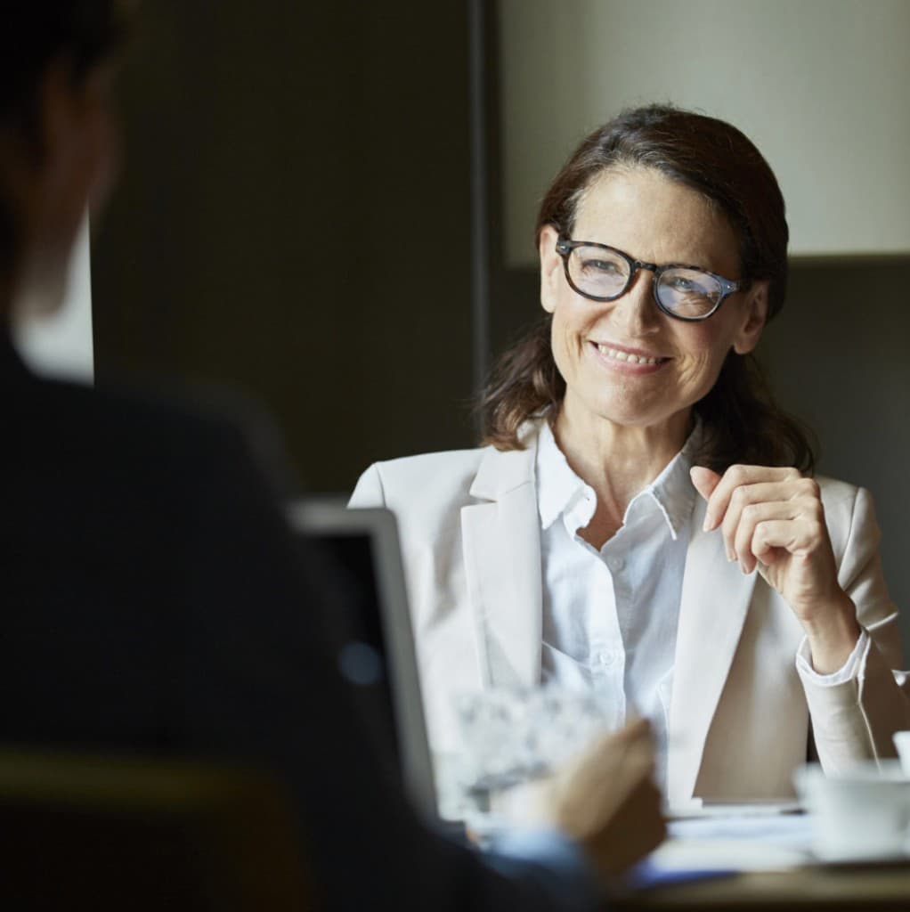 Seasoned advisor in conversation across a desk, laptop and coffee in soft focus—calm, direct counsel.