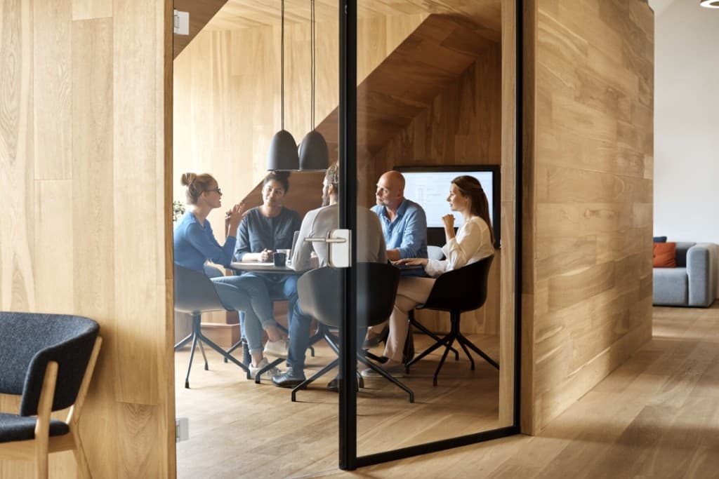 Team members engaged around a conference table with daylight and sheer curtains—deliberate, evidence-led dialogue.