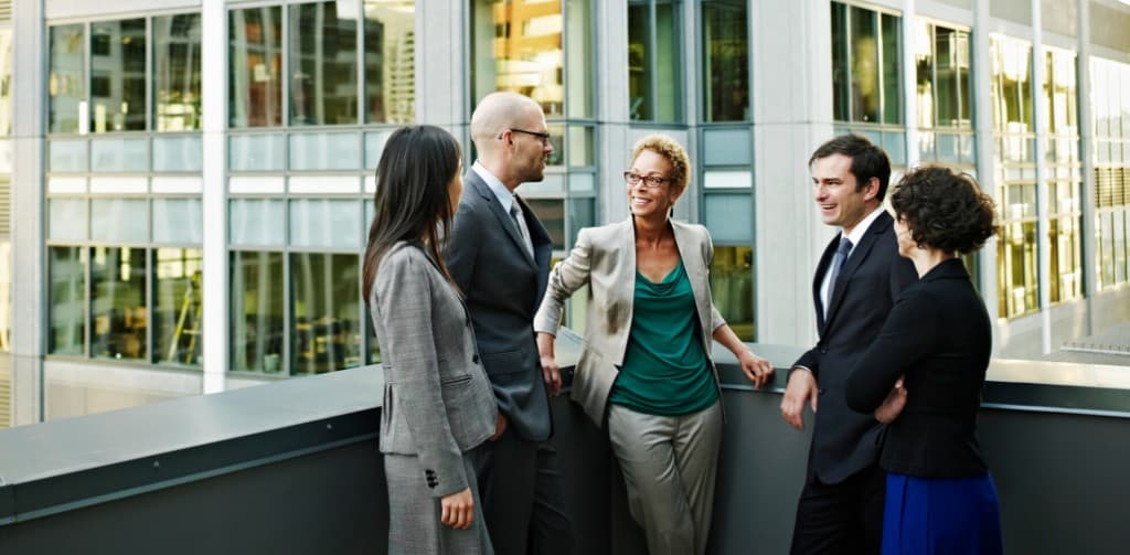 Small group of professionals in conversation on a terrace with city towers behind—institutional presence, human cadence.